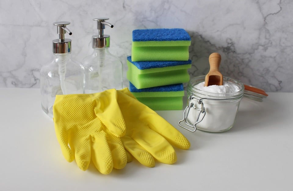 Un ensemble de produits de nettoyage sur une surface blanche comprend deux distributeurs de savon, plusieurs éponges vertes et bleues, une paire de gants en caoutchouc jaune et un pot en verre rempli de poudre blanche, probablement du bicarbonate de soude pour un entretien efficace, accompagné d'une petite cuillère en bois.